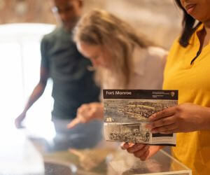 Three people inside the Casemate Museum. A close-up focuses on one holding the brochure for Fort Monroe. The others are in the background looking at exhibits.