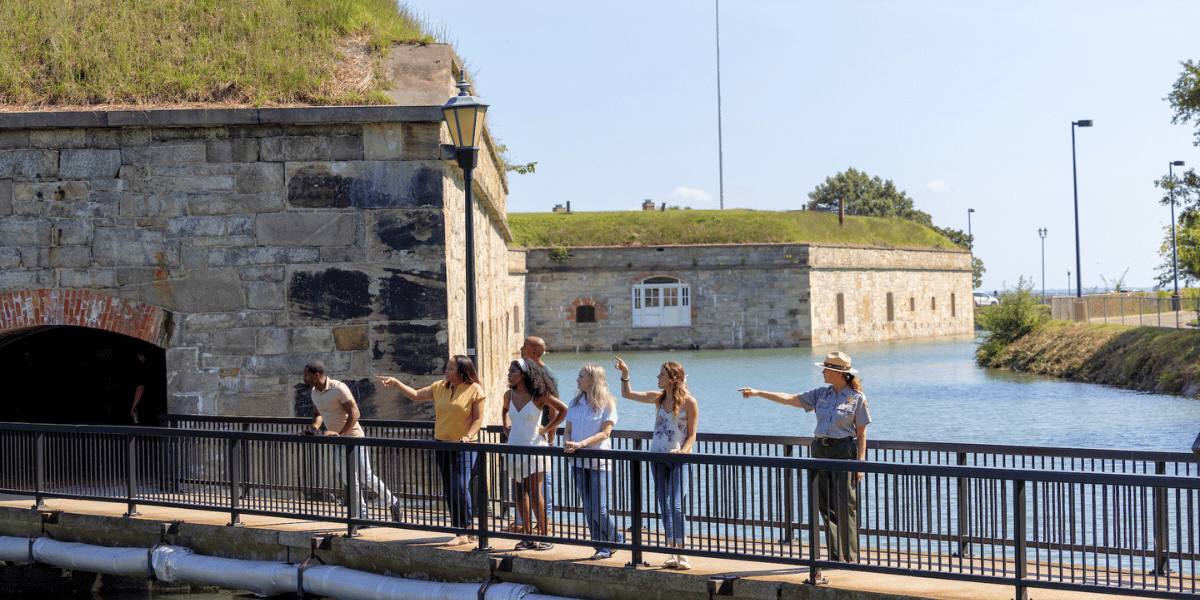 A park ranger leads a group of people on a bridge tour near a historic stone fort and moat. The scene is sunny, conveying a sense of exploration.