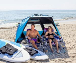 Couple relaxing on beach chairs under a beach tent with paddle boards beside them and beach in background.