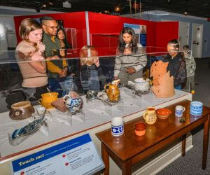 Family observe ceramic artifacts in a museum exhibit. Next to it, a table of modern recreations made from the same materials are available to pick up and hold. The scene is interactive and contemplative.
