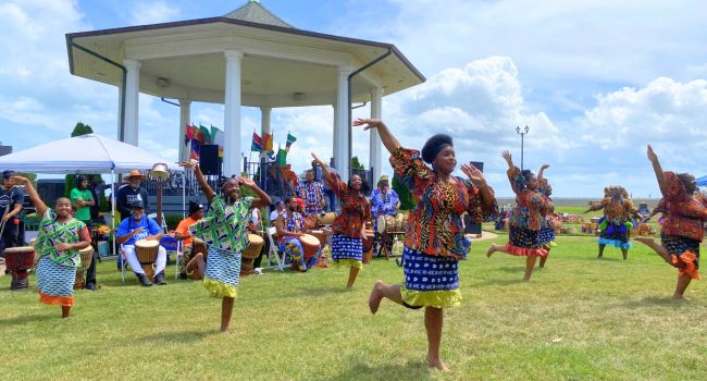 Women in colorful, traditional African garb dancing energetically on grass. Musicians play drums under a gazebo behind them.