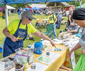 Volunteer in a bright Bee City apron at their outdoor market booth explains to an event attendee about composting using several props and flyers.