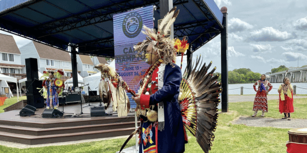 A person in traditional Native American attire performs a dance near a waterfront stage, displaying vibrant feathers and beadwork. Others in similar attire play instruments on stage. 