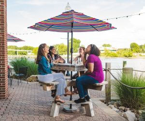 Four women dine outside under colorful umbrella with waterway in background.